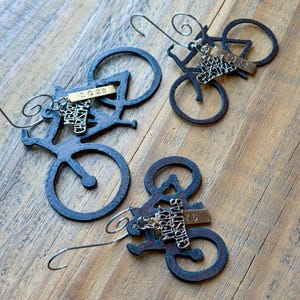 May include: Three dark brown metal bicycle ornaments, each featuring a silver-toned charm with the text Merry Christmas and a small rectangular brass-colored tag engraved with the year 2026, suspended by a curved metal wire hook for hanging.