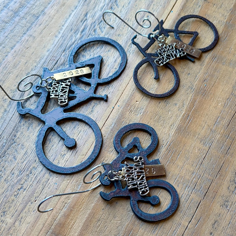 May include: Three dark brown metal bicycle ornaments, each featuring a silver-toned charm with the text Merry Christmas and a small rectangular brass-colored tag engraved with the year 2026, suspended by a curved metal wire hook for hanging.