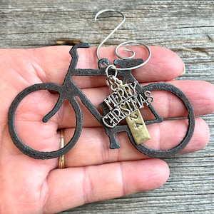 May include: A dark, textured metal bicycle ornament featuring a silver-toned wire hook. Attached to the frame is a silver-colored charm with the text MERRY CHRISTMAS and a small, rectangular brass-toned tag engraved with the number 24.