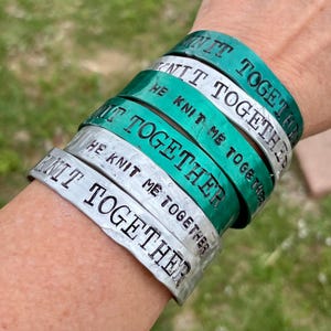 May include: A stack of silver and teal metal bracelets with the words "knit together" and "he knit me together" stamped on them. The bracelets are stacked on a wrist.
