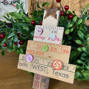 May include: A wooden Christmas tree ornament made from repurposed rulers. The tree is decorated with buttons, a star, and a red and white circle with the number 53. The ornament has the text "Country Store", "682-659", "North (WEST), Texas" on it.