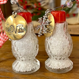 May include: Two clear glass salt and pepper shakers with red plastic caps, filled with white beads. Each shaker features a wire hanger with a clear bead, a gold-toned metal disc stamped with the word LOVE, and a silver-toned metal snowflake charm.