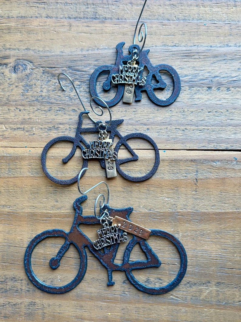 May include: Three dark metal bicycle ornaments with silver-toned wire hooks. Each bicycle features a dangling charm with the text Merry Christmas and a small rectangular metal tag engraved with the year 2026, suspended from the frame.