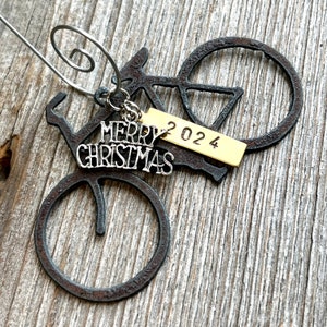 May include: A dark, rusted metal bicycle ornament featuring a silver-toned charm with the text Merry Christmas and a rectangular gold-toned metal tag stamped with the year 2024, all attached to a silver-toned wire spiral hanger.