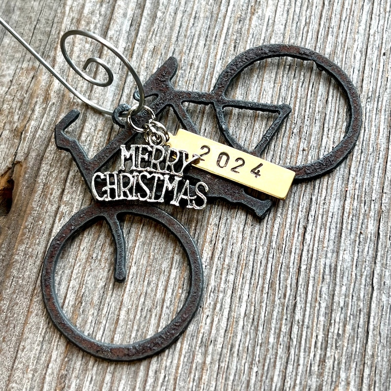 May include: A dark, rusted metal bicycle ornament featuring a silver-toned charm with the text Merry Christmas and a rectangular gold-toned metal tag stamped with the year 2024, all attached to a silver-toned wire spiral hanger.