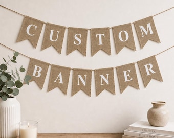 May include: A two-row burlap banner hanging on a string, featuring swallowtail flags with white letters spelling out CUSTOM BANNER. The banner is made of a textured, light brown fabric with clean-cut edges and bold, uppercase white lettering.