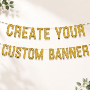 May include: A two-line banner featuring gold glitter letters strung on a thin cord. The top line spells out CREATE YOUR and the bottom line spells out CUSTOM BANNER. The letters are in a bold, sans-serif font with a sparkling, textured finish.