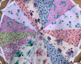 May include: A bunting with 14 triangular flags made of different floral patterned fabrics in shades of pink, blue, green, and white. The flags are sewn together with white fabric and have white ties.