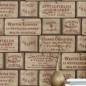 May include: Interior shot with a light yellow chest of drawers, a pink armchair, and a framed botanical print. The wallpaper mimics wooden crates, displaying text like "Finest Olive Oil" and "Weston Lobster". The room has a vintage aesthetic.