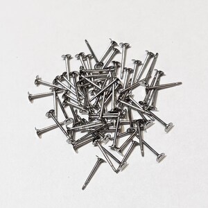 May include: A collection of small, silver-coloured metal nails with flat, circular heads and thin, pointed shafts. These fasteners are arranged in a loose, overlapping pile, showcasing their metallic finish and uniform, slender construction.