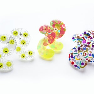 May include: Three clear plastic hair clips with different designs. The first clip has a pattern of yellow smiley faces on white daisies. The second clip has a pink and yellow swirl pattern. The third clip has a colourful confetti pattern.