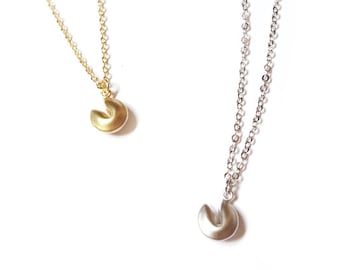May include: Two necklaces featuring small fortune cookie-shaped pendants. One necklace is gold-toned with a delicate chain, and the other is silver-toned with a matching chain. Both pendants have a smooth, matte finish and a curved, folded design.