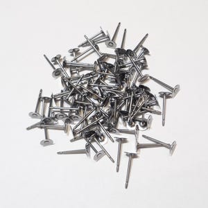 May include: A collection of numerous silver-toned metal earring posts, each featuring a flat circular pad at the top for attaching components. These small, slender jewellery findings are arranged in a loose, overlapping pile on a plain surface.