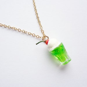 May include: A gold chain necklace with a green and white miniature ice cream soda charm. The charm has a red cherry on top.
