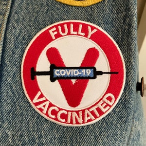 May include: A red and white circular patch with the text "FULLY VACCINATED" and a stylized red "V" with a blue syringe inside. The syringe has the text "COVID-19" on it.