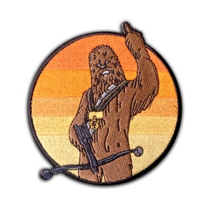 May include: Embroidered patch featuring Chewbacca from Star Wars holding a bowcaster in front of a sunset. The patch is circular and has a black outline.