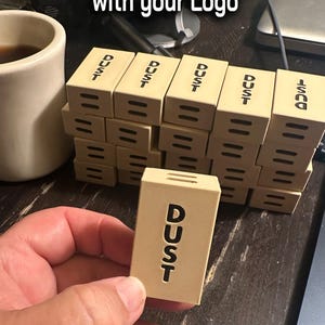 Könnte beinhalten: Eine Sammlung von 3D-gedruckten Mini-Appleboxen in hellem Beige, einige gestapelt und andere in einer Hand gehalten. Jede Box trägt das Wort "DUST". Der Text "3D Printed Mini Mini Applebox with your Logo" befindet sich oben.