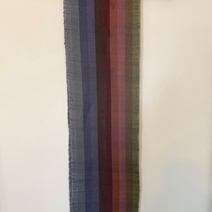 Op de afbeelding: Een geweven textiel dat aan een houten roede hangt. Het textiel heeft verticale strepen in tinten grijs, blauw, paars, rood en groen. De onderste rand heeft een franje van bijpassende kleuren. De roede is donkerbruin.