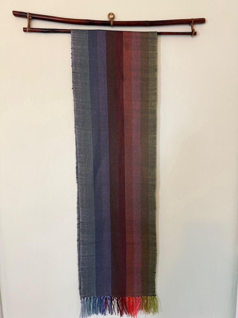 Op de afbeelding: Een geweven textiel dat aan een houten roede hangt. Het textiel heeft verticale strepen in tinten grijs, blauw, paars, rood en groen. De onderste rand heeft een franje van bijpassende kleuren. De roede is donkerbruin.