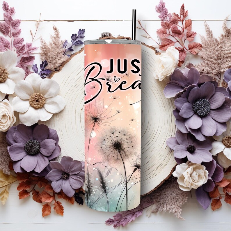 Op de afbeelding: Een roestvrijstalen beker met een zwart rietje. De beker heeft een paardenbloemontwerp met de woorden "Just Breathe" in zwart schrift. De achtergrond heeft een verloop van roze, perzik en teal kleuren.
