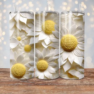 May include: Three white stainless steel tumblers with a pattern of white paper flowers with yellow centers. The tumblers are on a wooden surface.
