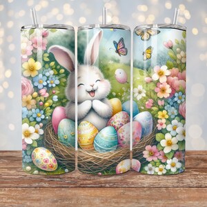 May include: A white bunny with pink ears sits in a nest of twigs with colorful Easter eggs. The bunny is smiling and has its paws clasped together. The background is a green field with pink and blue flowers.