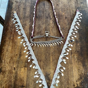 Puede incluir: Un conjunto decorativo de cinturón y collar. El collar es de color púrpura oscuro con cuentas blancas y una cadena plateada. El cinturón presenta una cadena plateada con piezas metálicas decorativas. El collar y el cinturón tienen adornos de encaje blanco con adornos plateados.