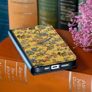 Op de afbeelding: Een zwarte telefoonhoes met een geel bloemenpatroon met vogels, bloemen en takken. De hoes is open en onthult de rand en poorten van de telefoon. Het ontwerp bevat roze, blauwe en groene accenten. De hoes ligt op een bruin boek.