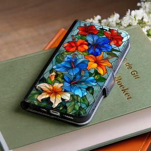 May include: A black phone case featuring a colorful floral design with red, blue, and orange hibiscus flowers. The case has a leather-like texture and a magnetic closure. The design resembles stained glass.