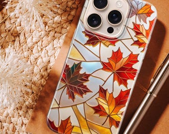 May include: A clear phone case with a stained-glass effect, depicting autumn maple leaves in red, orange, and yellow. The case has a glossy finish and a protective bumper. The phone case is shown on a brown surface with a pen.