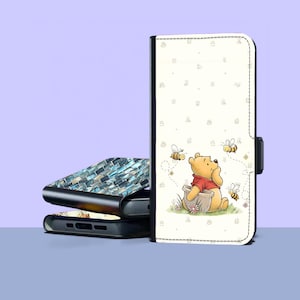 Puede incluir: Una funda de teléfono con una ilustración de dibujos animados de Winnie the Pooh sentado en una olla, rodeado de abejas. La funda es blanca con un borde negro y un cierre magnético. Otras fundas con un diseño de mosaico azul y blanco están apiladas detrás.
