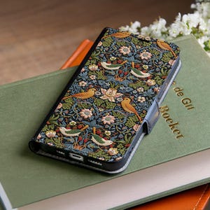 Puede incluir: Una funda de teléfono negra con un colorido estampado floral y de pájaros. El diseño presenta pájaros, flores y bayas en tonos amarillos, verdes y rosas sobre un fondo oscuro. La funda está abierta, revelando la pantalla del teléfono.