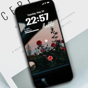 May include: A black smartphone displays a floral wallpaper with red roses and a full moon. The time is 22:57, and the word "hello" is written in cursive. The phone rests on a white surface with the word "CEREAL" visible.