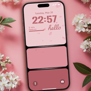 以下が含まれることがあります： 22:57と「hello」の文字が表示された、ピンクと赤のグラデーションのロック画面を持つスマートフォン。白い花に囲まれ、ピンク色の表面に置かれています。画面には日付の5月20日（火）も表示されています。