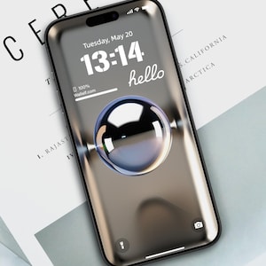 May include: A black smartphone with a metallic finish displays a clock face with the time 13:14 and the word "hello" on the screen. The date is Tuesday, May 20. The phone is on a white surface.