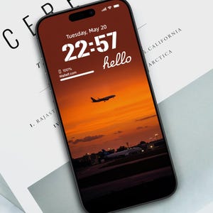 May include: A smartphone displays a sunset scene with an airplane taking off. The screen shows the time 22:57, the word "hello", and the text "100% Walletf.com". The phone is black and rests on a white surface with the word "CEREAL" visible.