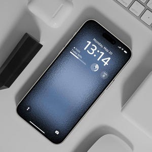 Puede incluir: Un teléfono inteligente negro con una pantalla texturizada que muestra la hora 13:14 y la fecha lunes 20 de mayo. El teléfono está sobre una superficie blanca con un teclado blanco y un ratón blanco.