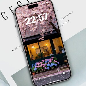 May include: A smartphone shows a floral wallpaper with a pink cherry blossom tree and a shop front. The time is 22:57 on Tuesday, May 20. The phone rests on a white surface with the word "CEREAL" visible.