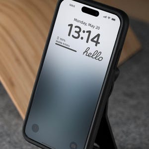 以下が含まれることがあります： 黒いスマートフォンで、画面には13:14の時間、5月20日月曜日の日付、そして「hello」という言葉が表示されています。スマートフォンは黒いケースに入っていて、黒いスタンドの上に置かれています。