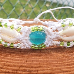May include: White macrame bracelet featuring a central turquoise bead, flanked by two seashells. The bracelet is adorned with green and clear beads. The design is set against a natural wood surface, suggesting a beach or summer theme.