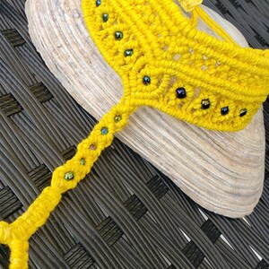 May include: A vibrant yellow macrame anklet, adorned with small, colourful beads. The anklet is presented on a seashell, highlighting its intricate woven pattern. The anklet's bright colour and detailed craftsmanship make it a unique accessory.