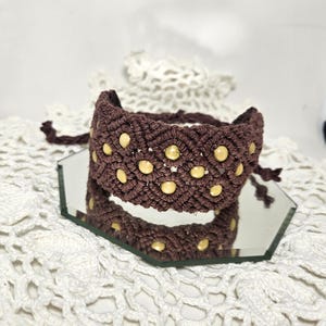 May include: A dark brown macrame bracelet featuring a woven diamond pattern with embedded round, light-colored beads. The bracelet has knotted cord ends for fastening and is displayed on a hexagonal mirror surface.