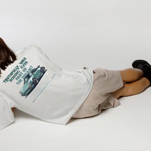 May include: White t-shirt with a green car graphic and text that reads "Truthfully was boring ever part of the dream?". The person is wearing beige shorts and black boots.