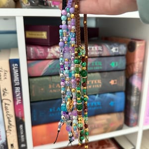 Op de afbeelding: Een hand houdt meerdere kralenarmbanden in verschillende kleuren vast, waaronder paars, groen en goud. De armbanden zijn geregen op koorden en hebben verschillende maten en vormen kralen. De achtergrond is een witte plank vol boeken.