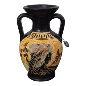 Peut inclure: Un vase en céramique noir et beige avec deux anses et une large ouverture. Le vase présente un motif peint d'un profil de femme et des motifs décoratifs. Une petite louche en métal est suspendue sur le côté.