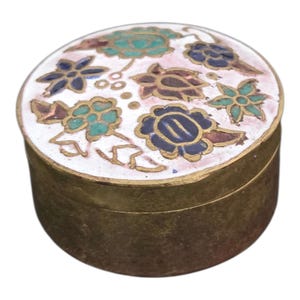 May include: A small, round trinket box with a white lid featuring a floral design in blue, teal, and brown. The box has a gold-colored rim and base. The floral pattern is set against a pink and white background.