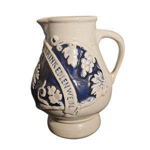 May include: Cream ceramic pitcher with a handle and spout. A blue emblem with grape and leaf designs is on the front, with the words "TRINK EDLEN WEIN" in white. The pitcher has a rounded base and a capacity marking.