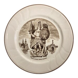 May include: White ceramic plate with a brown rim and a detailed sepia-toned illustration of Saint Michael. The image depicts Saint Michael with a sword and shield, standing over a defeated figure. Text at the bottom reads "SANCTE MICHAEL REPELLE DRAGONES."
