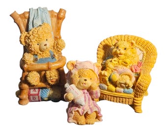 Puede incluir: Tres figuras decorativas de osos de peluche. Un oso está sentado en una silla de bambú, otro sostiene un biberón y lleva un gorro de dormir, y el tercero está en una silla de mimbre con una manta. Los osos son amarillos y visten ropa azul y rosa.