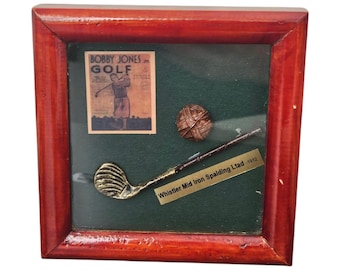 May include: A framed display featuring a vintage magazine cover of Bobby Jones on Golf, a miniature golf club, a golf ball, and a gold plaque that reads "Whistler Mid Iron Spalding Ltd 1912".